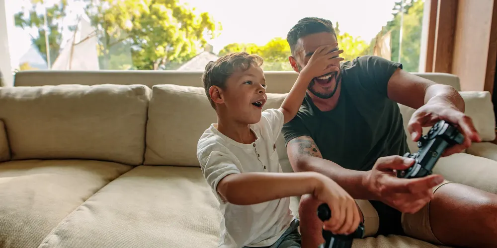 Little boy covering eyes of his father playing video game. Cheerful family of father and son having fun playing video games at home. Courtesy Jacob Lund/Shutterstock