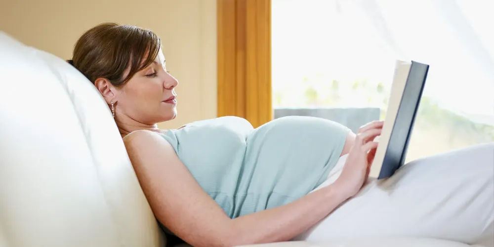 7 months pregnant woman lying down on sofa and reading book.; Courtesy Diego Cervo/Shutterstock
