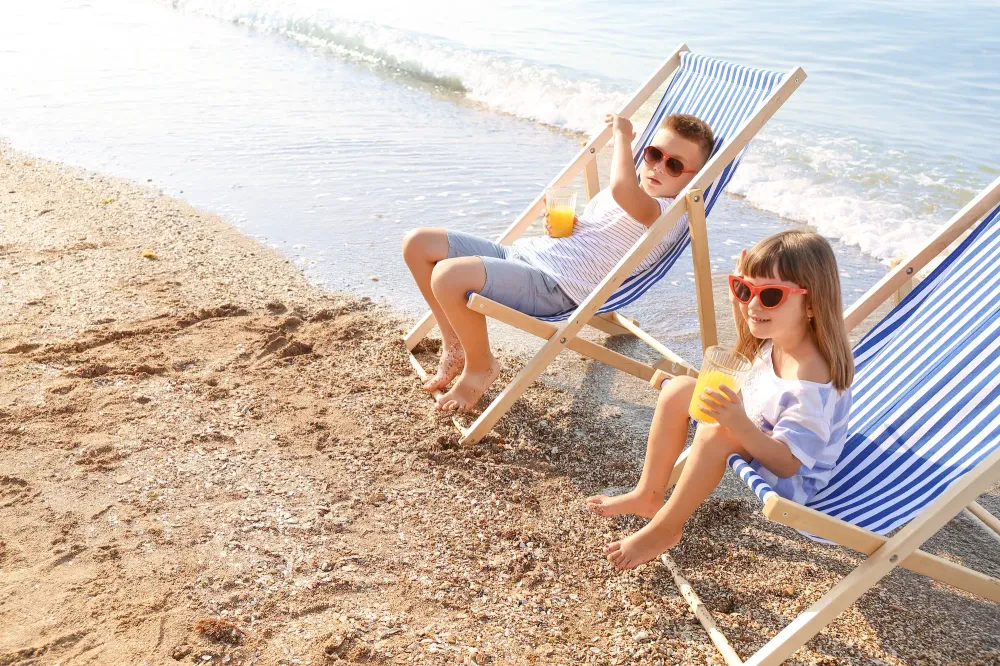Two children drinking juice on the beach in beach chairs