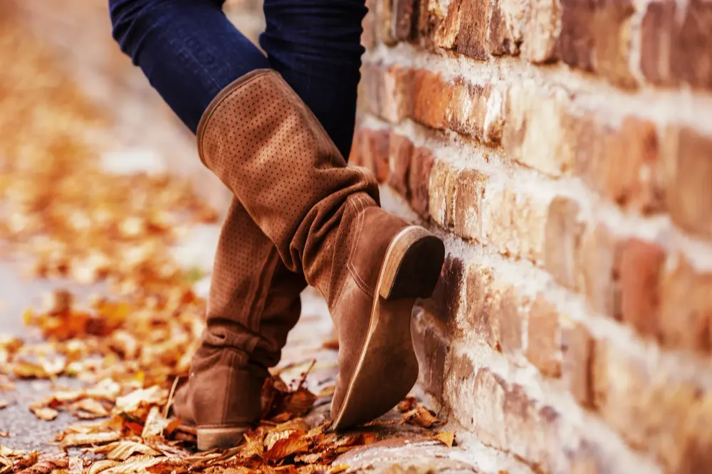 Person wearing tall fall boots, leaning against a brick wall and standing on fallen leaves