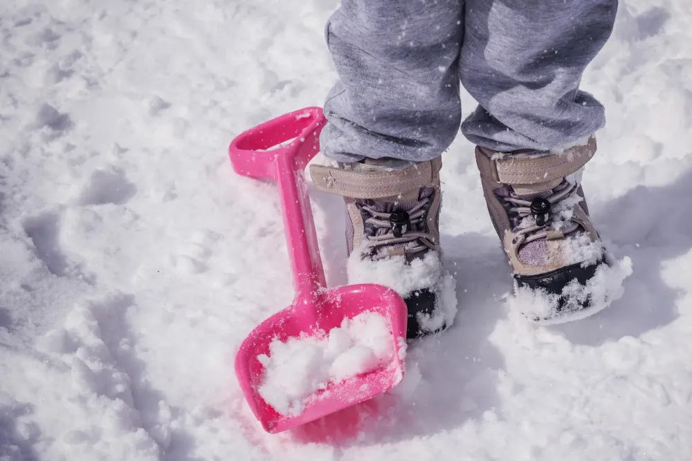 Child wearing snow boots in the snow next to a snow toy shovel