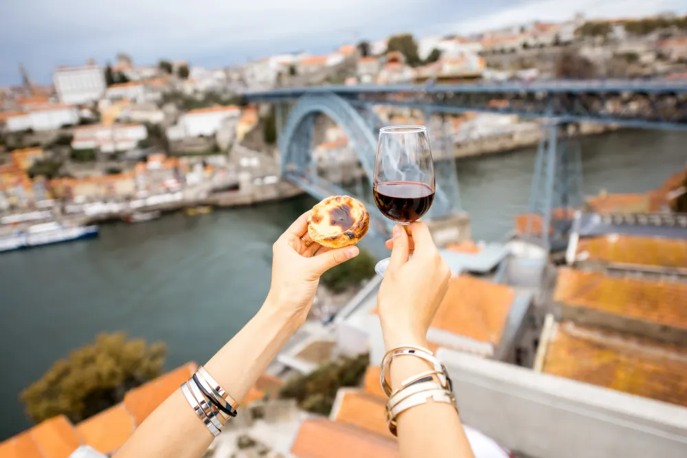 Hands holding glass of port wine & pastel de Nata on cityscape background of Porto, Portugal